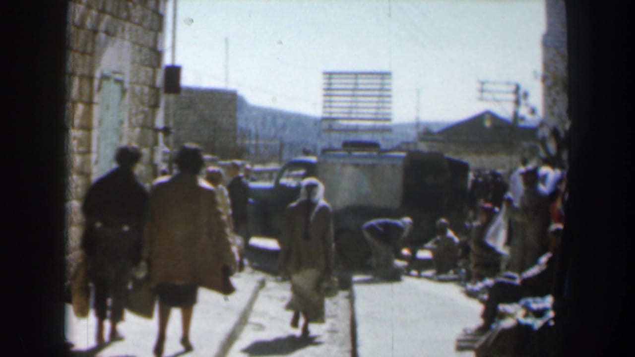 Pedestrians walk along a narrow sunlit street beside stone walls and a dark vehicle.