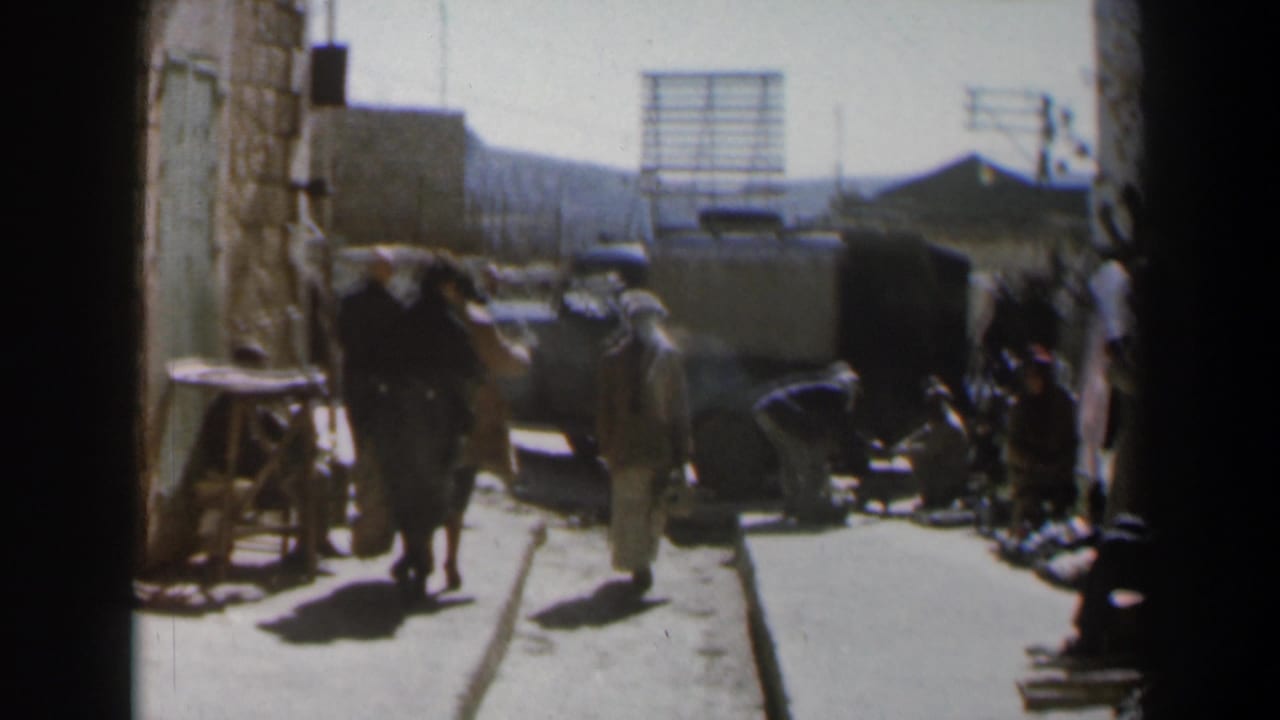 People move along the same street near a dark vehicle and stone building wall.
