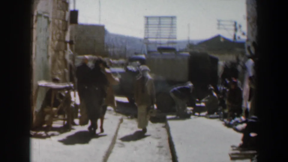 1960 Israel street frame with pedestrians, seated figures, a dark vehicle, stone walls, and strong midday sunlight.