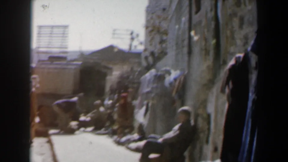 1960 Israel home-movie frame showing a narrow street lined with stone walls, seated people, pedestrians, and a vehicle at left.
