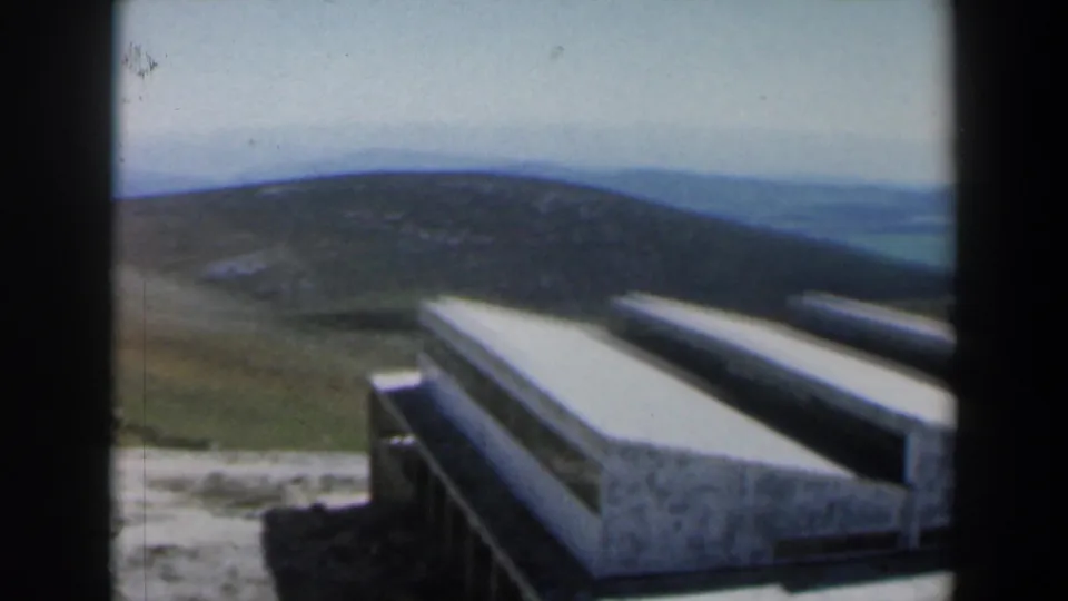 1960 Israel home-movie frame looking from the street area toward a green hill and distant landscape beyond pale foreground structures.