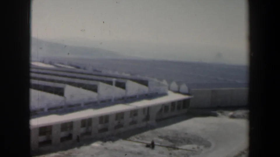 Wider 8mm view of the Israeli hillside site, showing the concrete structure, a building edge, open ground, and hazy hills.