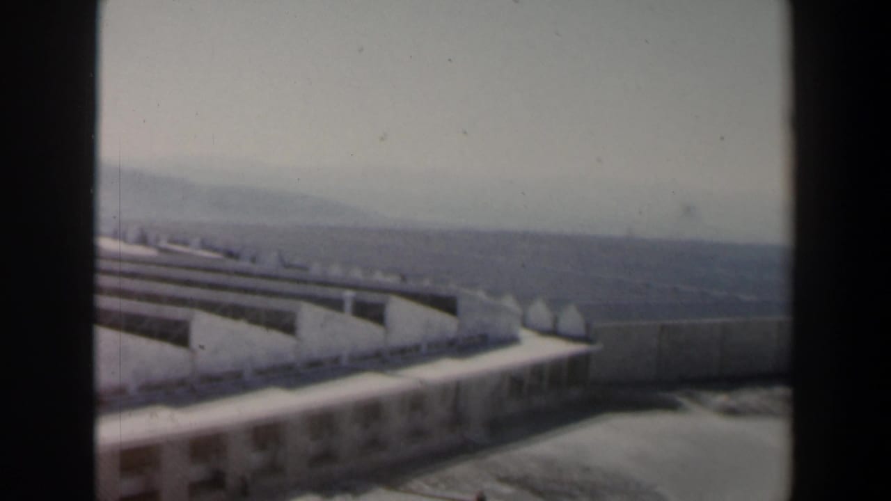1960 Israel 8mm frame showing a large modernist building complex with pale roofs, concrete walls, and hazy hills beyond.