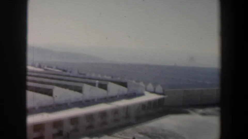 1960 Israel 8mm frame showing a large modernist building complex with pale roofs, concrete walls, and hazy hills beyond.