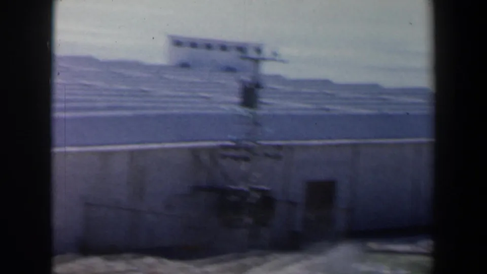 1960 Israel 8mm frame showing a blurred concrete compound exterior with ribbed surfaces, a low wall, and hills beyond.
