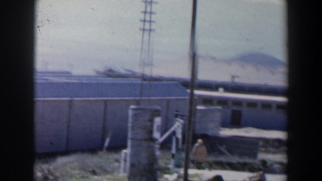 Concrete compound seen through dark vertical edges, with utility poles, walls, and distant hills.