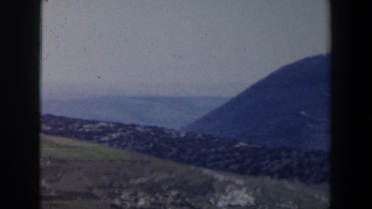 Aerial 8mm view over an Israeli coastal hillside settlement with water and a rounded mountain beyond.