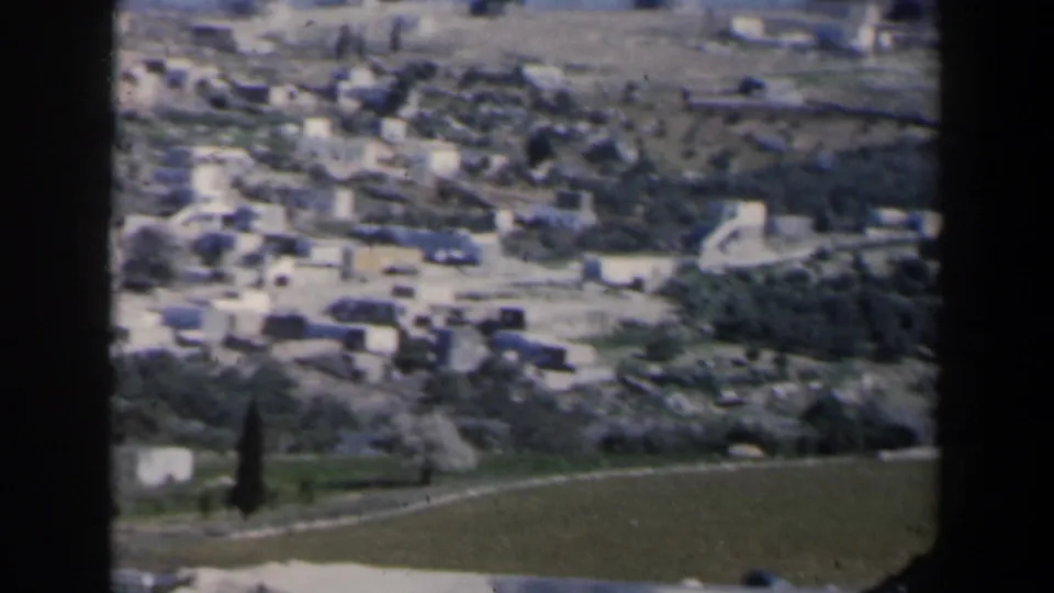 Aerial 8mm view of a small Israeli town with light buildings, roads, fields and hills nearby.
