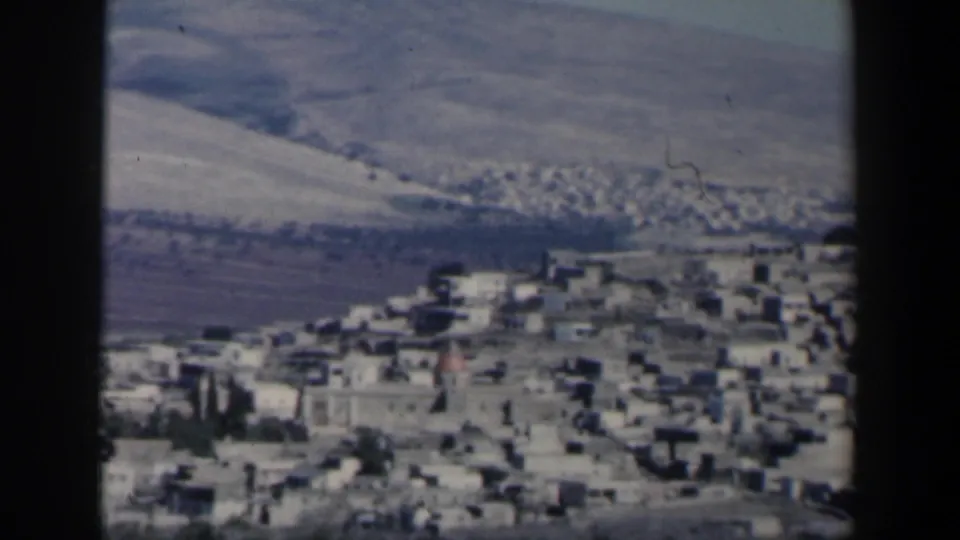 1960 Israel 8mm frame showing a dense hillside town, pale rooftops, sparse vegetation, and dry terrain beyond.