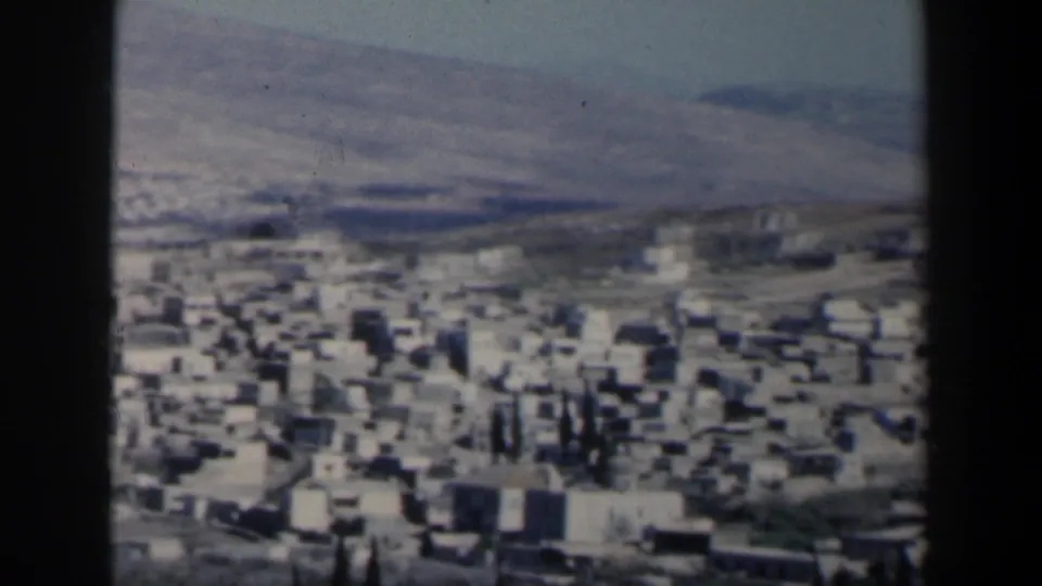 1960 8mm view over a hillside town in Israel, with pale buildings, sparse trees, roads, and distant hills under a bright sky.