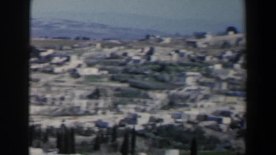 Soft 8mm frame of an Israel hillside settlement, with white structures, green patches, roads, and dark vertical trees in the foreground.