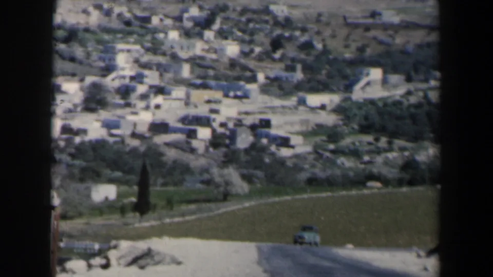 1960 Israel hillside town view with white buildings above fields, cypress-like trees, a curving road, and a small car near the lower edge.