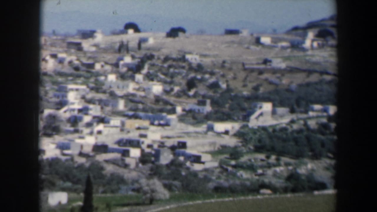 Wide 8mm travel view in Israel showing pale buildings spread across a hillside above fields and scattered trees.