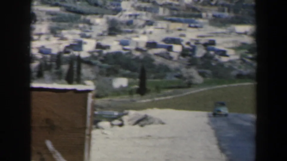 Israel home-movie frame showing a small car on a road below fields, dark trees, and pale hillside buildings.