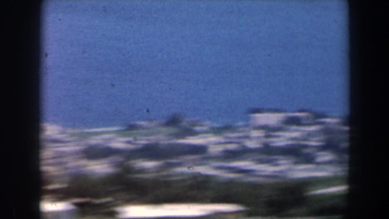 Soft 8mm frame showing a distant Israeli coastal townscape in 1960, with pale buildings, blue sky, and dark foreground borders.