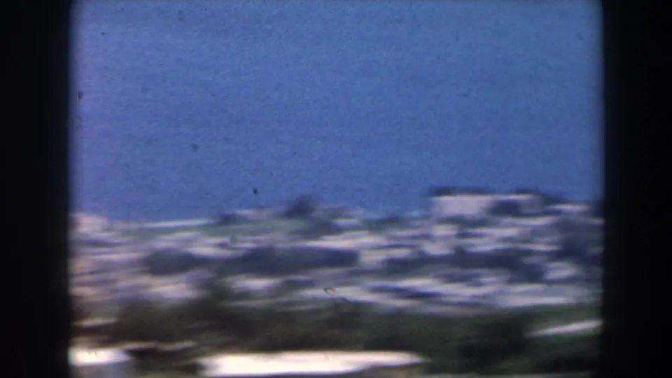 Soft 8mm frame showing a distant Israeli coastal townscape in 1960, with pale buildings, blue sky, and dark foreground borders.