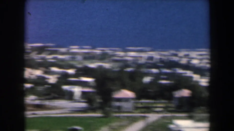 Elevated 1960 Israel coastal view with low pale buildings, open green land, distant sea, and soft faded 8mm color.