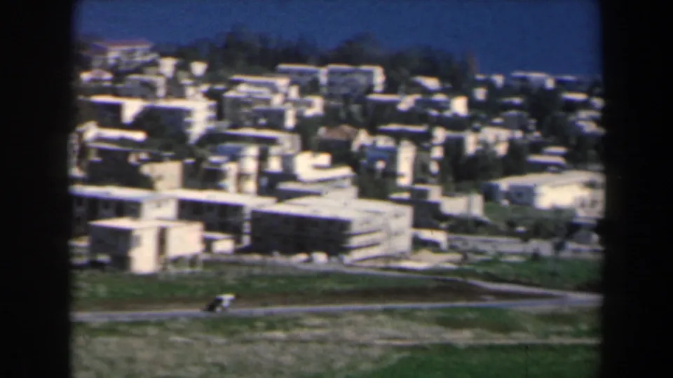Clearer 1960 Israeli town view with clustered white buildings, hillside roads, green foreground, and blue sky in an 8mm home-movie frame.
