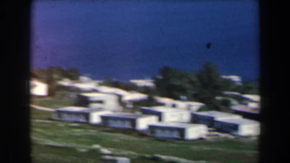 1960 Israel 8mm frame showing white residential blocks beyond open grass, paths, and trees in a soft home-movie view.