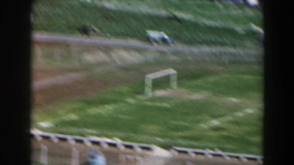 1960 Israel 8mm frame showing a green football field, goalpost, dirt track, trees, and distant residential buildings.