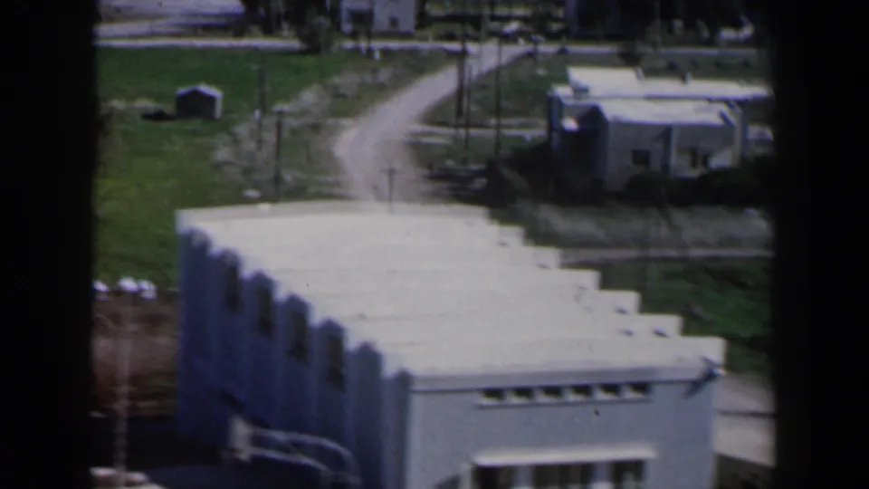 1960 Israel 8mm frame showing a white modernist building near green fields, utility poles, and unpaved roads.