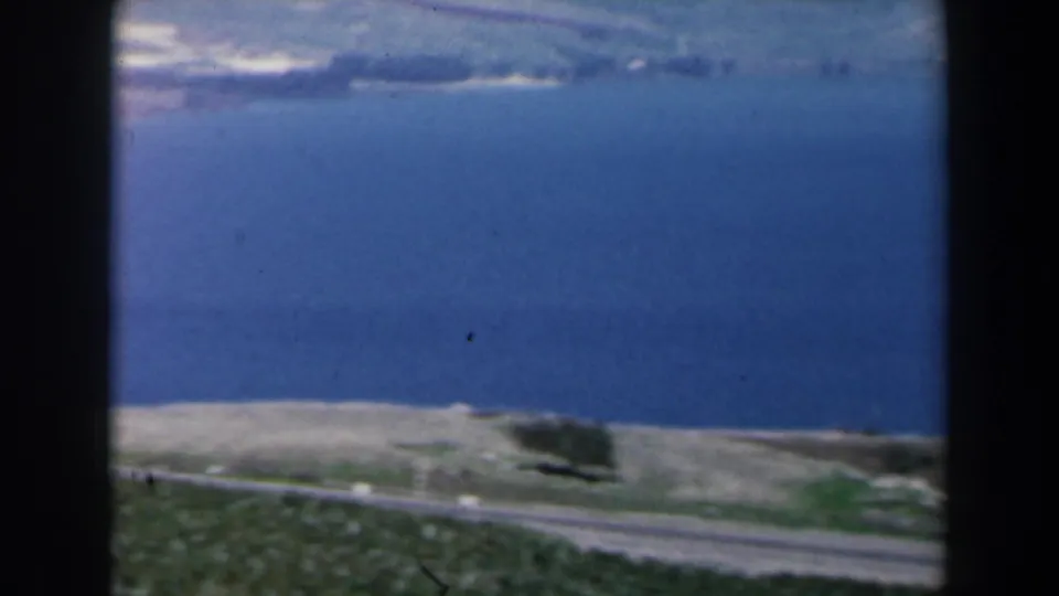 1960 Israel 8mm frame looking across blue water, a pale shoreline, roadside ground, and cultivated fields.