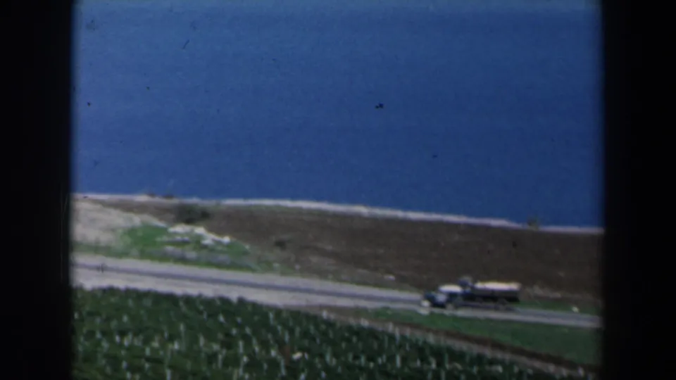 1960 Israel 8mm frame showing a small vintage truck on a coastal road beside blue water and agricultural plots.