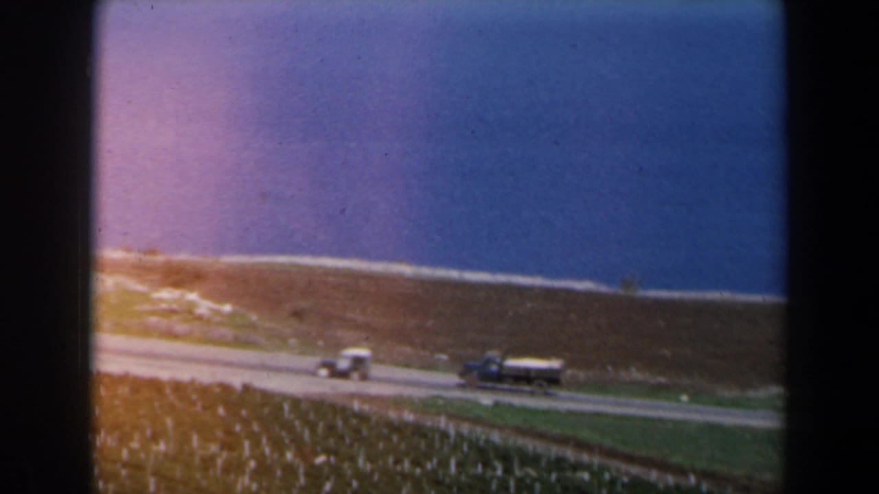 Coastal road above green fields with a small car and truck near a blue sea.