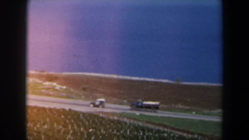 1960 Israel 8mm frame showing a road along the coast, a small car, a truck, green farmland, dry hillside, and blue water beyond.