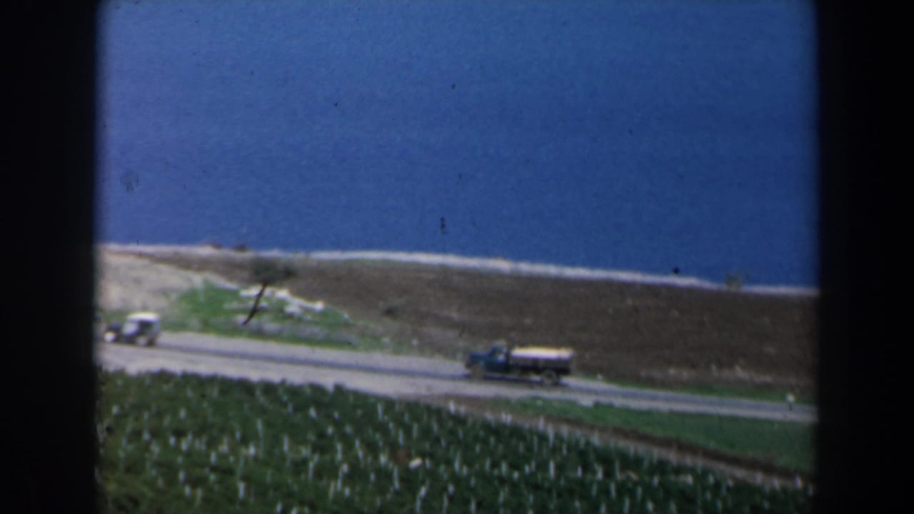 Truck and small car on a shoreline road with fields and blue water beyond.
