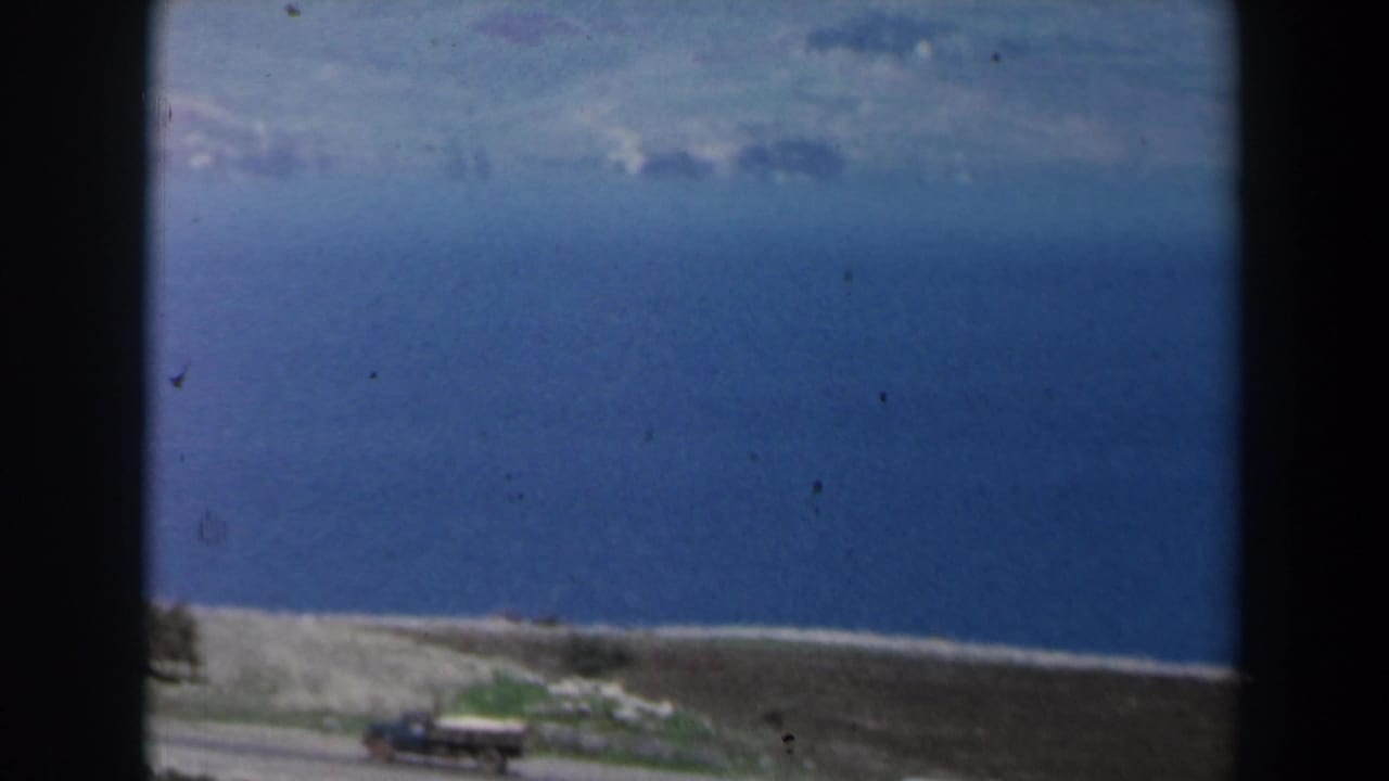 1960 Israel 8mm frame showing a lake, pale shoreline, road, and distant hills under a bright sky.