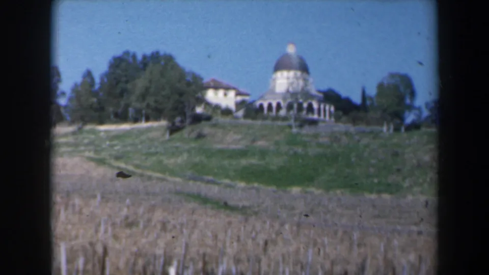 1960 Israel 8mm frame showing a distant white domed building on a hill, with dry grass, paths, and trees under a blue sky.