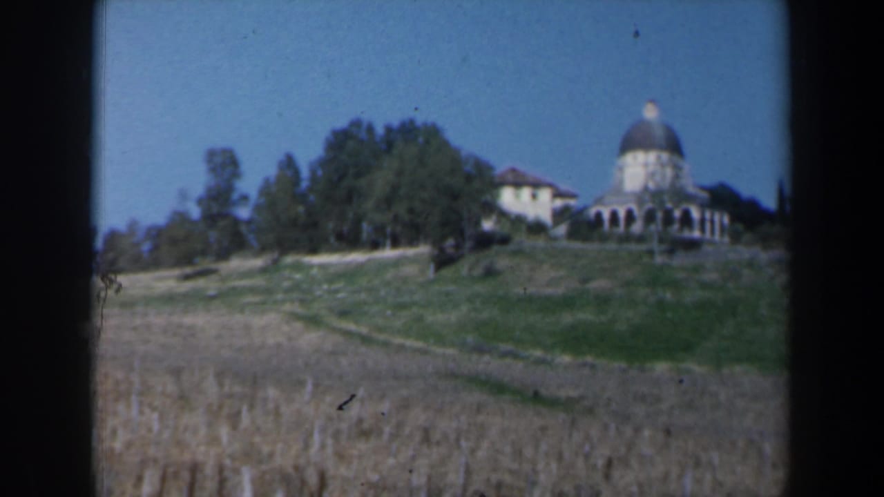 1960 Israel 8mm view of a domed structure beside a pale building, with trees, sloped grass, dry field, and clear sky.