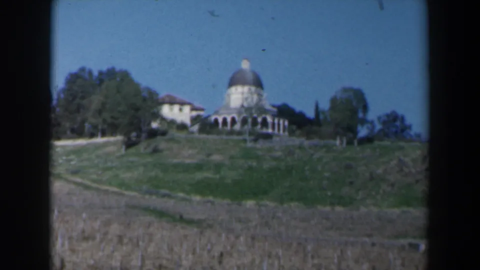 1960 Israel home-movie frame with a domed hillside building, neighboring pale structure, trees, grassy slope, and dry field below.