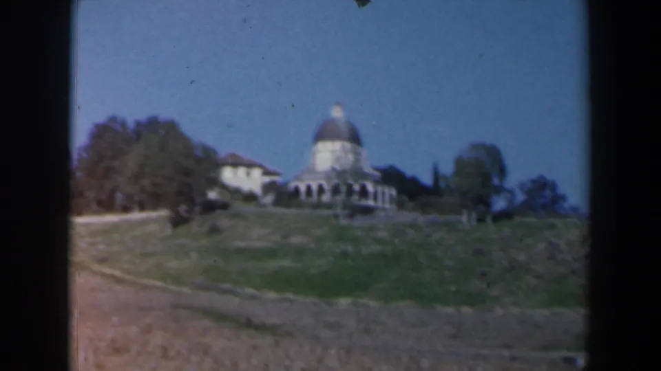 1960 Israel 8mm frame showing a wide view of the domed hillside building, wooded slope, open dry field, and blue sky.