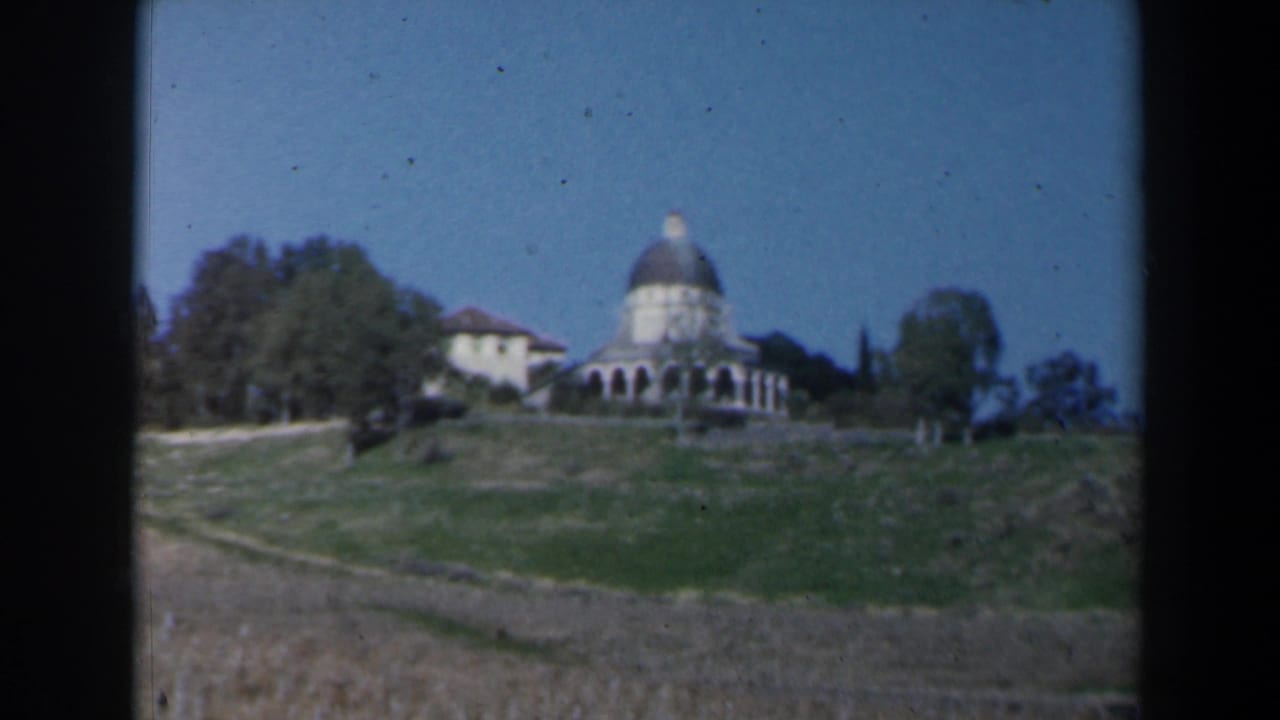 Domed building and adjacent structure on a grassy hill with trees and blue sky.