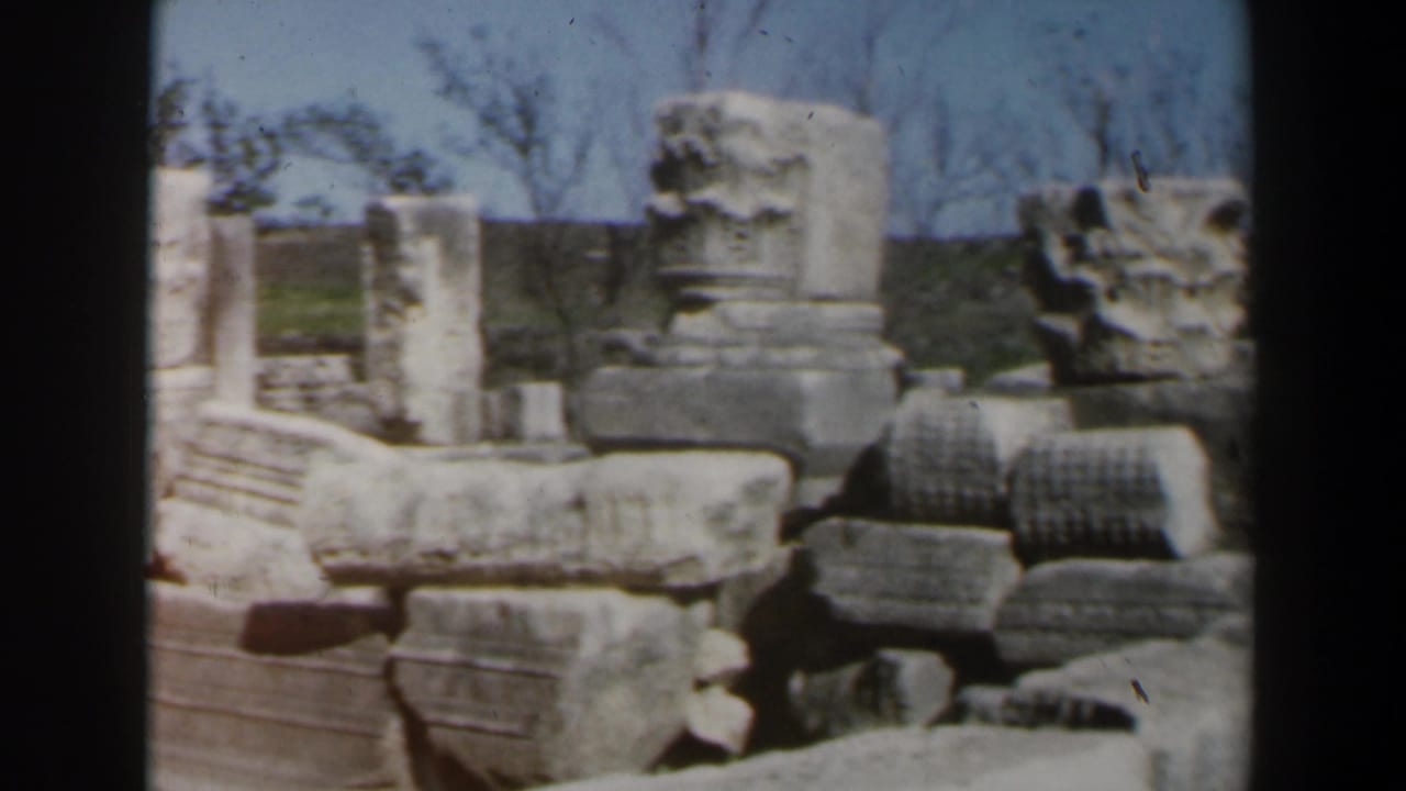 1960s Israel home-movie frame showing weathered stone blocks, column fragments, and low ruins at an open-air archaeological site.