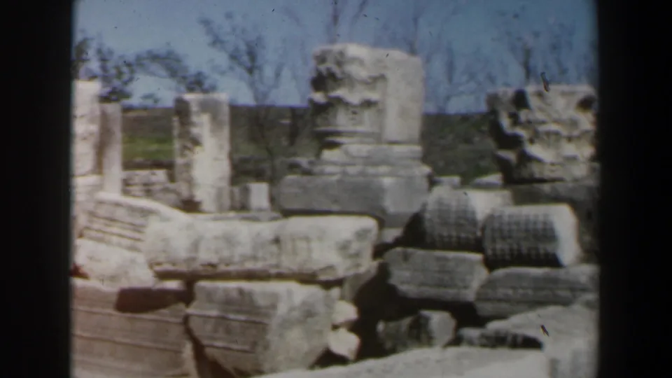 1960s Israel home-movie frame showing weathered stone blocks, column fragments, and low ruins at an open-air archaeological site.