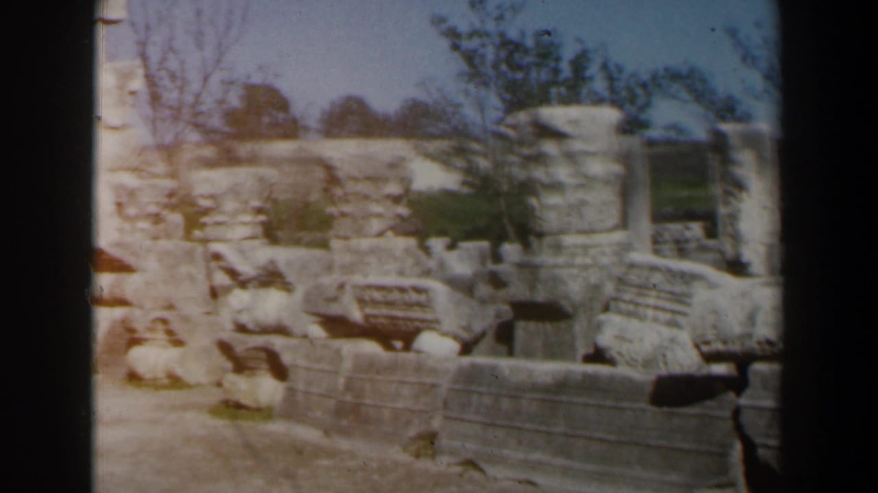 Open ruins with broken stonework, low walls, and a tree beyond the masonry.