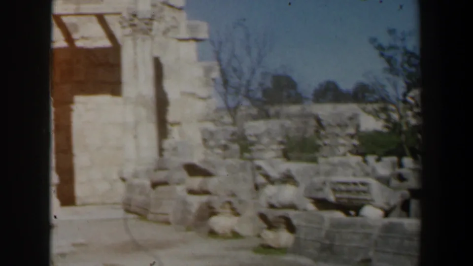1960s Israel home-movie frame showing a stone doorway or wall edge beside carved blocks and scattered archaeological remains.