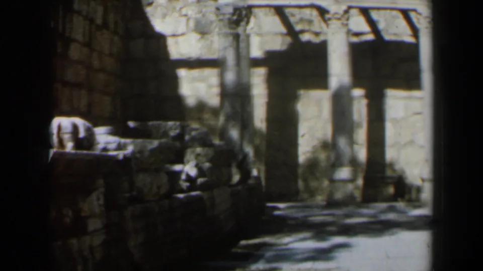 1960 Israel home-movie frame showing a low stone wall, columns, rubble, and deep shadows across an ancient ruin site.
