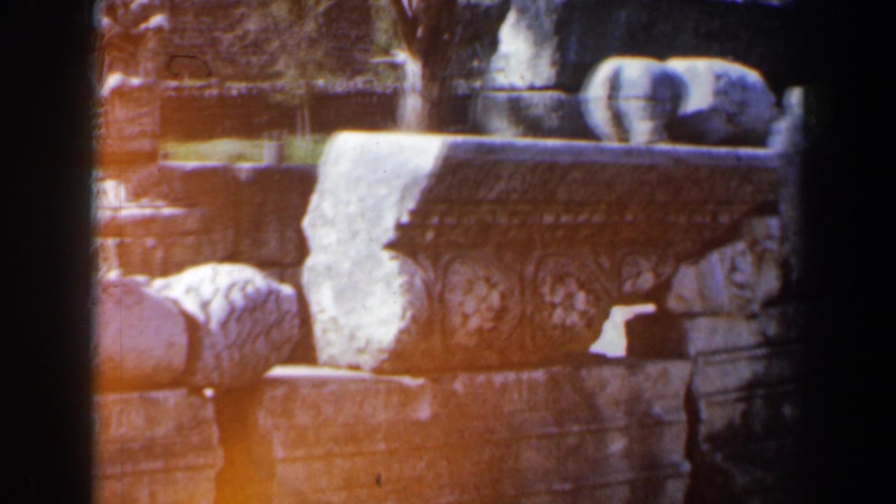 1960 8mm Israel travel frame showing stacked ancient stone fragments, a floral carved capital, and dark gate shadows.