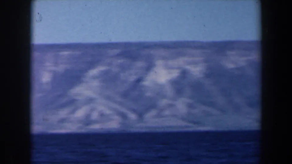 1960 8mm Israel home-movie frame showing sea in the foreground and a soft, rugged coastal landscape beyond.