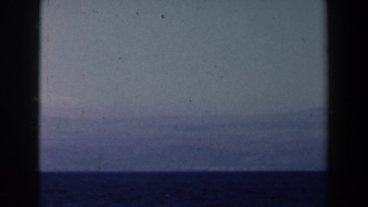 A distant hazy shoreline or mountain range appears above dark blue water in 1960 Israel-context 8mm footage.