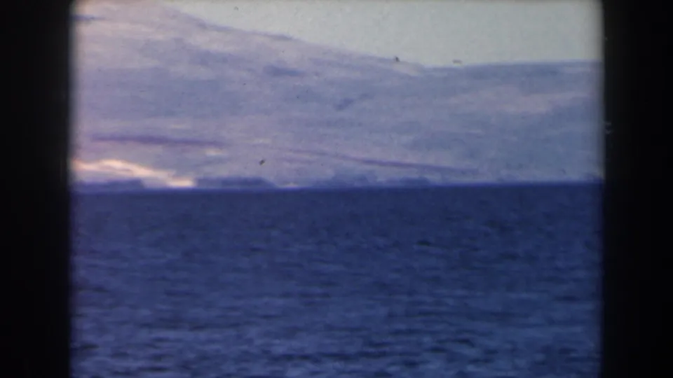 1960 Israel 8mm frame showing blue sea, a low bright shoreline, and soft mountain forms under a washed pale sky.