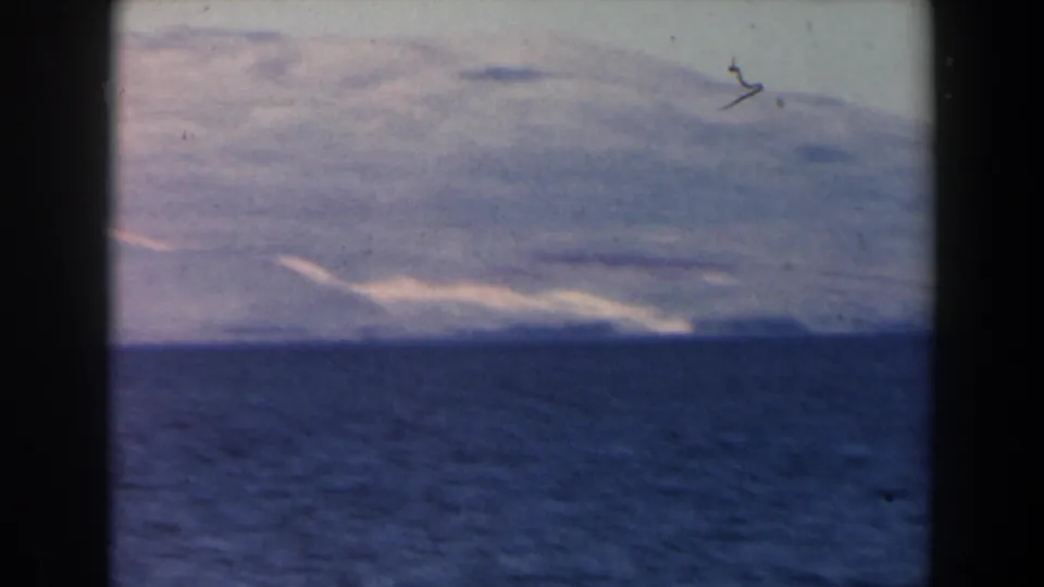 1960 Israel 8mm frame with dark water in the foreground, a faint surf line or light shoreline, and hazy mountains beyond.