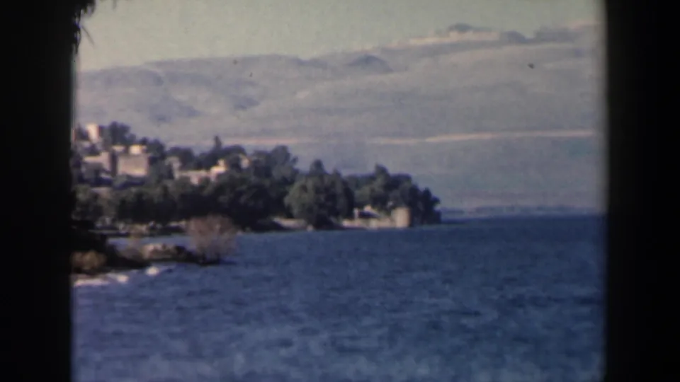 1960 Israel 8mm view of a tree-lined shoreline with small buildings, blue coastal water, and pale hills under a hazy sky.