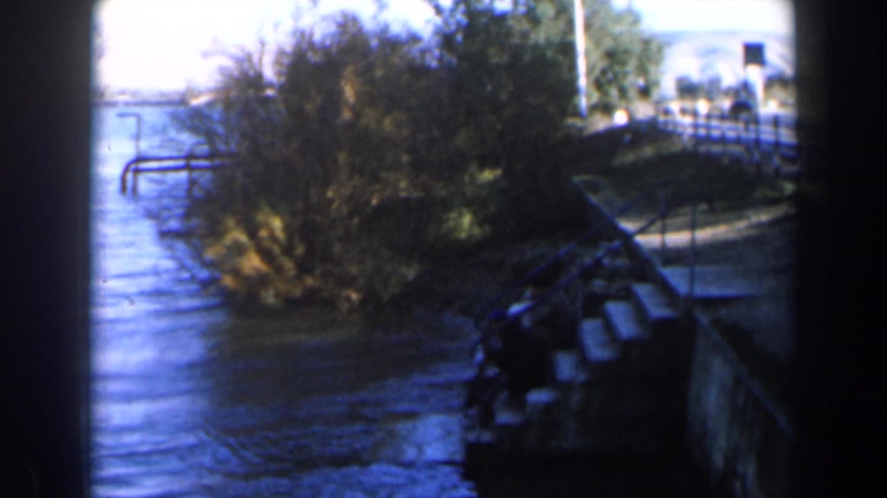 River beside concrete steps, brush, a road guardrail, and distant pale hills.