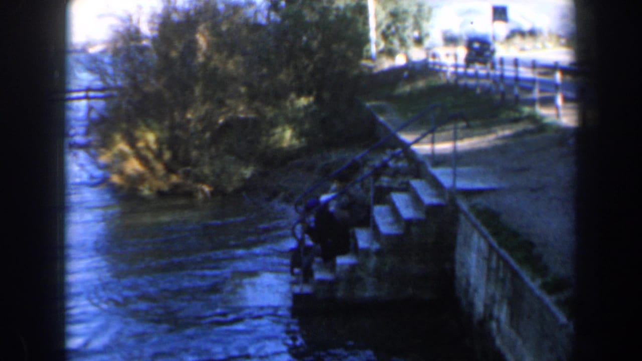 1960 Israel home-movie frame with water flowing past steps, shoreline brush, a road above, and mountains beyond the river corridor.