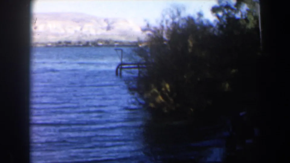1960 Israel 8mm frame centered on the river surface, bank vegetation, and pale mountain slopes beyond the water.
